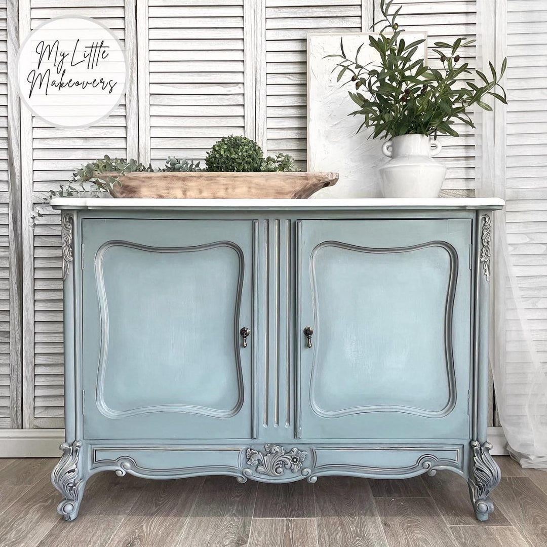 A distressed light blue and silver French-style cabinet with a white marble top, styled with a wooden tray and potted greenery against a white shutter backdrop.