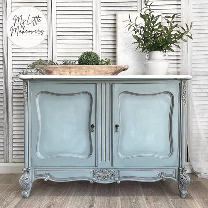 A distressed light blue and silver French-style cabinet with a white marble top, styled with a wooden tray and potted greenery against a white shutter backdrop.