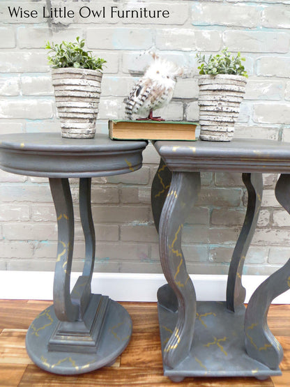 A pair of round console tables with distressed gray painted wood and gold crackle detailing, styled with potted plants and a small owl figurine against a faux brick wall.