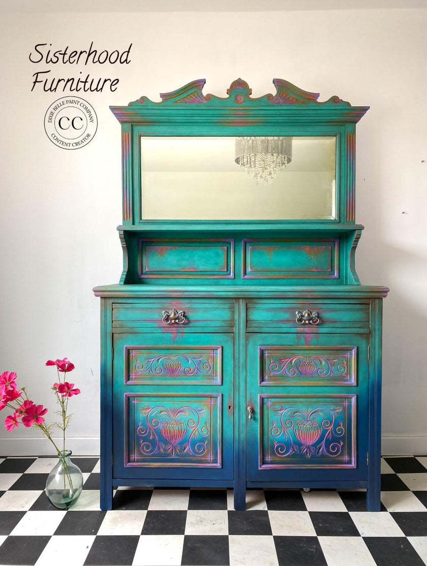 A turquoise and blue distressed wooden hutch cabinet with a mirror on top, set against a white wall on a black-and-white checkered floor. Pink flowers in a glass vase sit to the left.