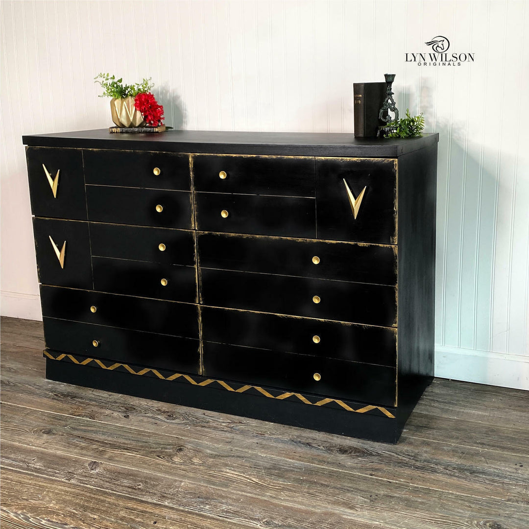 A black wooden dresser with gold accents and multiple drawers, styled in a room setting with decorative items on top, a visible brand watermark in the upper right, and a light paneled wall and wood floor background.