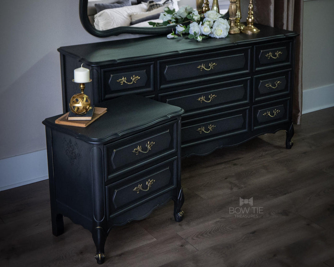 A dark painted wooden dresser with a matching small side table, styled in a bedroom setting with a mirror, flowers, and table lamps on a wood floor.