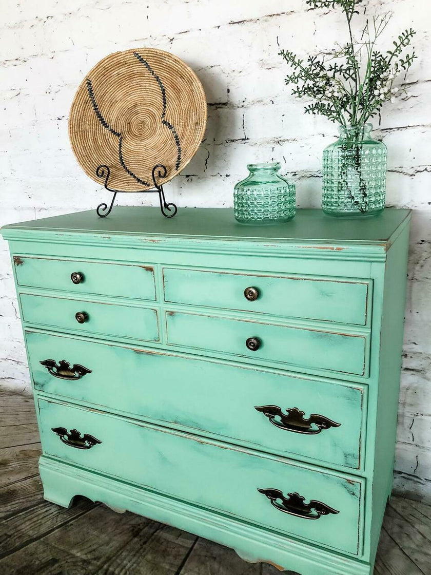 A distressed mint green wooden dresser with dark metal drawer pulls, styled with a woven plate on a stand and two glass vases against a white brick wall.