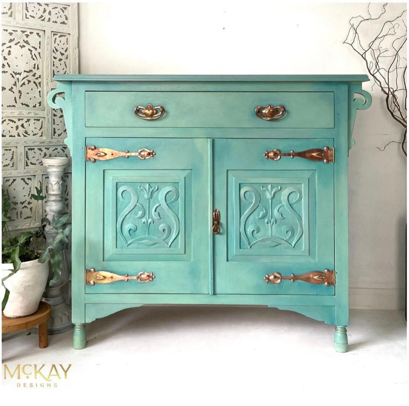 A turquoise painted wooden sideboard cabinet with ornate carved door panels and antique brass handles, shown in a styled interior setting with decorative wall panels and plants.