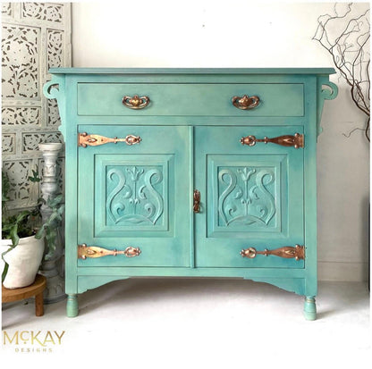 A turquoise painted wooden sideboard cabinet with ornate carved door panels and antique brass handles, shown in a styled interior setting with decorative wall panels and plants.