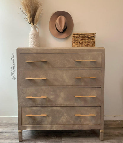 A rustic, distressed gray-brown wooden chest of five drawers with light wood handles, styled in a home setting with a hat, wicker basket, and dried grass in a vase on top.