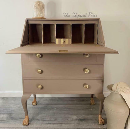 A distressed taupe-brown wooden secretary desk with claw legs and decorative brass knobs, shown against a plain wall with a grey wood floor. A small vase sits on top, and a large ceramic pot is on the right.