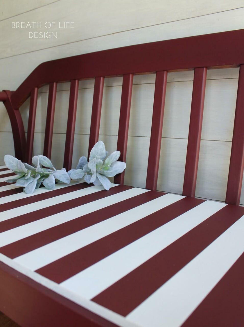 A close-up of a painted wooden bench or chair backrest with alternating dark red and white painted slats, set against a light wood paneled wall. Two white artificial flowers rest on the seat.