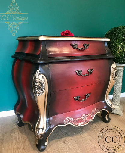 A vintage-style three-drawer chest of drawers with a glossy dark red and black finish, ornate gold-painted cabriole legs, and dark metal drawer pulls, set against a teal wall.