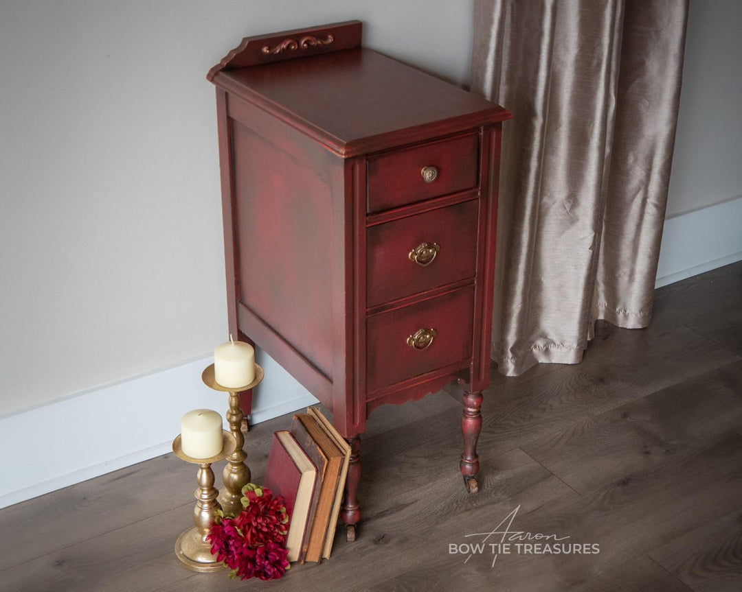 A small vintage-style reddish-brown wooden nightstand with three drawers and brass handles, styled in a room corner with books, candles, flowers, and a curtain.