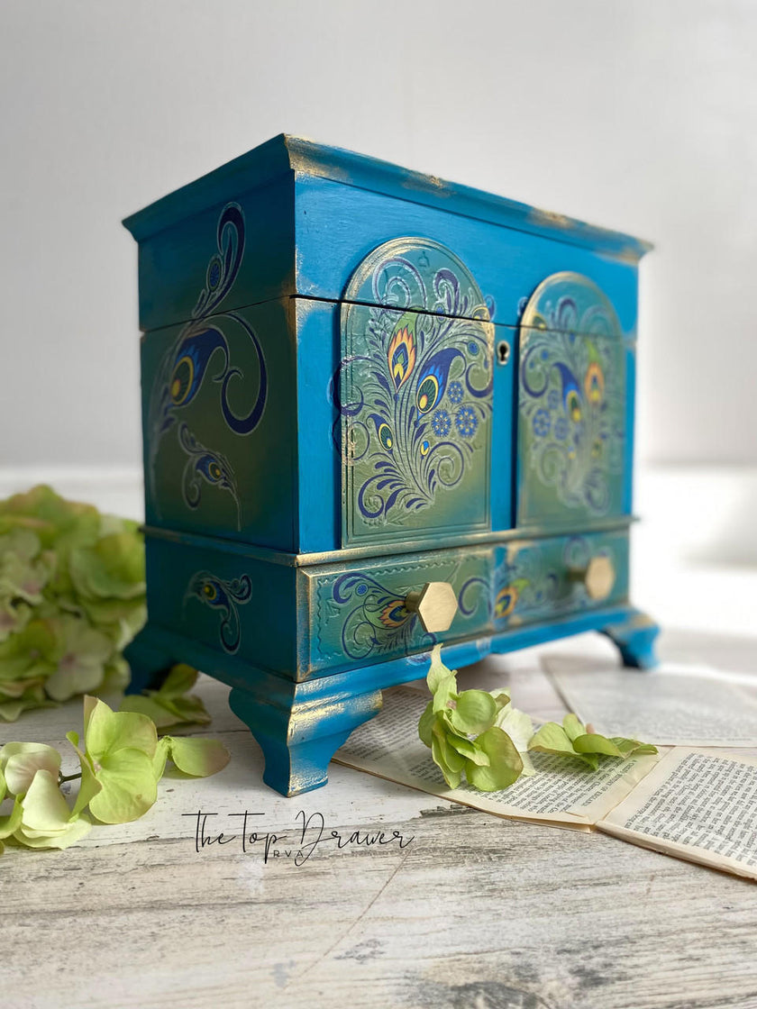 A turquoise blue decorative jewelry box with hand-painted peacock and floral motifs, styled on a rustic wooden surface with green hydrangea petals and an open book.