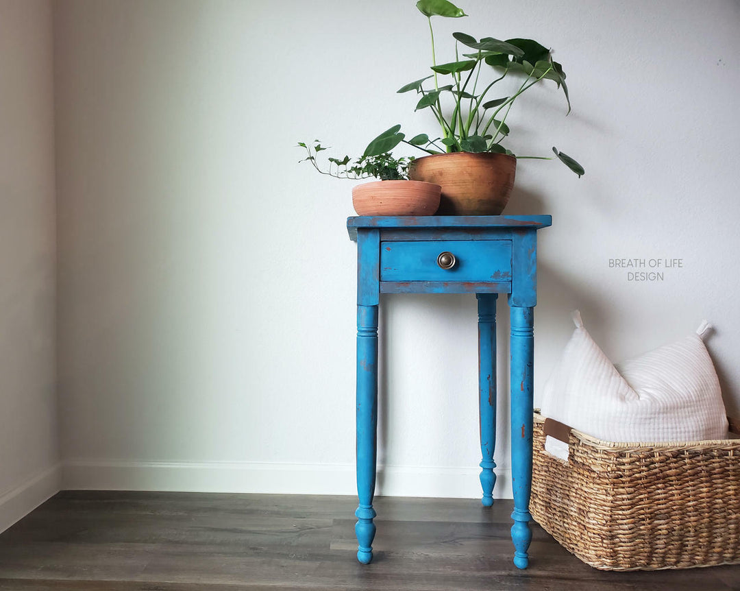 A distressed blue wooden single-drawer side table with turned legs, styled with potted plants on top and next to a woven basket with a white pillow in a minimalist room setting.