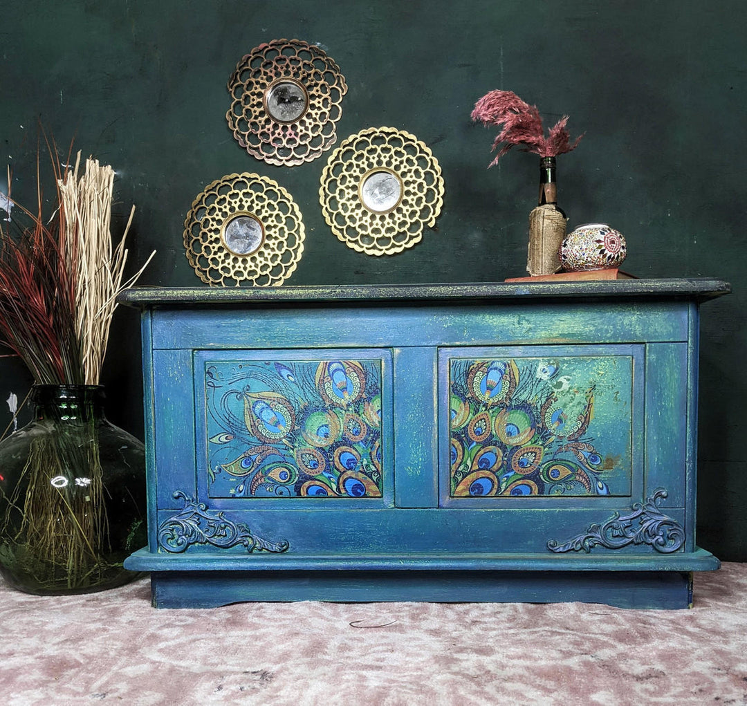 A distressed teal blue wooden sideboard cabinet with peacock feather painted details on the front doors, styled with decorative gold plates, a vase of dried pink grass, and a patterned bowl against a dark green wall.