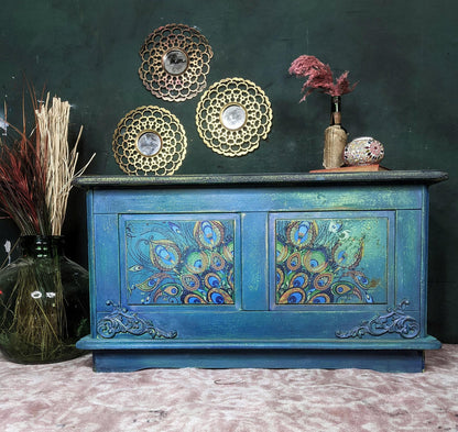 A distressed teal blue wooden sideboard cabinet with peacock feather painted details on the front doors, styled with decorative gold plates, a vase of dried pink grass, and a patterned bowl against a dark green wall.