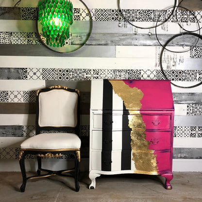 A painted three-drawer chest of drawers with a bold vertical color-blocked design in white, black, and bright pink, accented with gold leaf on the right side. It is styled in a room setting next to a matching black and cream chair, with a patterned wall and hanging glass ornaments in the background.