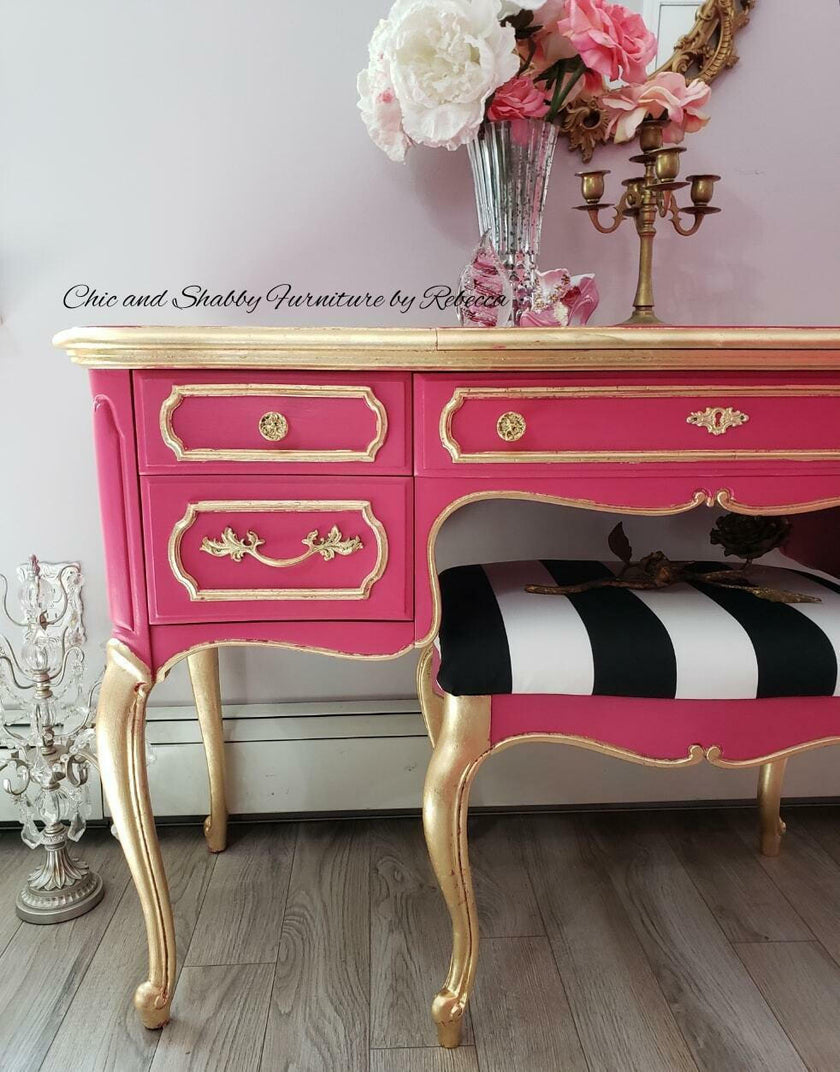A pink and gold French-style writing desk with a matching striped upholstered bench, styled in a home interior setting with a vase of flowers and a gold candelabra on top.
