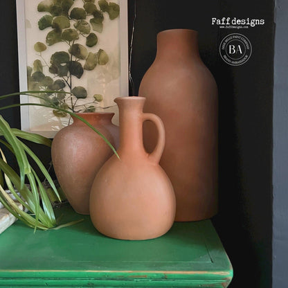 Three matte terracotta clay vases in different shapes displayed on a distressed green shelf against a dark background, with a framed botanical print and a plant leaf visible.