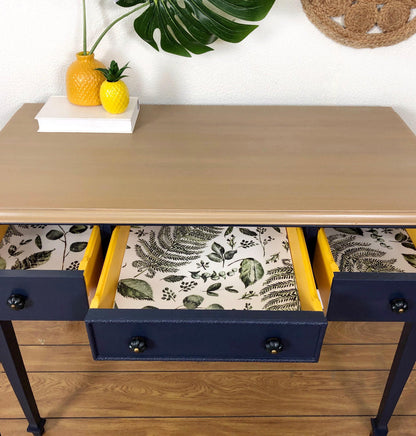 A navy blue writing desk with a light wood top, featuring three open drawers with botanical patterned linens and bright yellow drawer interiors. The desk is styled with a vase, book, and pineapple on a white surface, set against a white wall with a green plant leaf and a woven wall hanging.