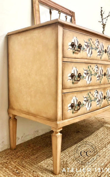 A vintage-style three-drawer dresser in a warm beige and gold finish, decorated with white floral motifs and ornate dark metal handles, shown in a rustic room setting.