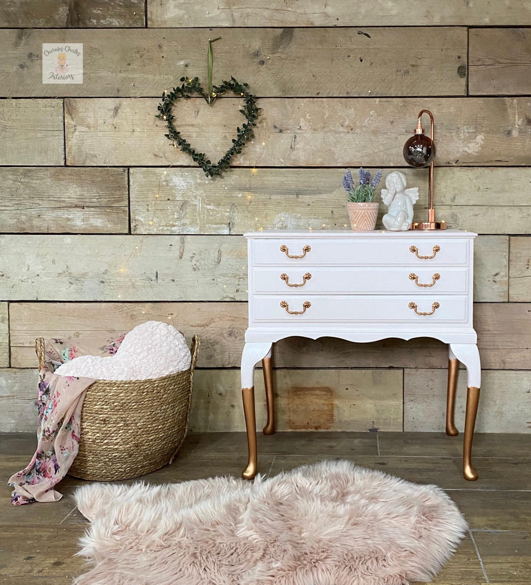 A white three-drawer dresser with gold-tone handles and legs, styled in a rustic room setting against a wood plank wall with a pink faux fur rug, a woven basket, and a heart-shaped greenery wall hanging.