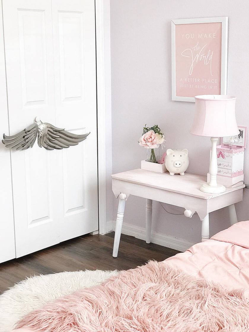 A styled bedroom corner with a pale pink bedspread, fluffy pink throw blanket, white nightstand, pink lamp, framed wall art, and a white door with gray angel wings mounted on it.