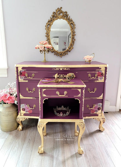 A purple and gold vintage-style vanity table with floral hand-painted details, styled with a matching gold mirror, flowers, and a tiara on a light wood floor.