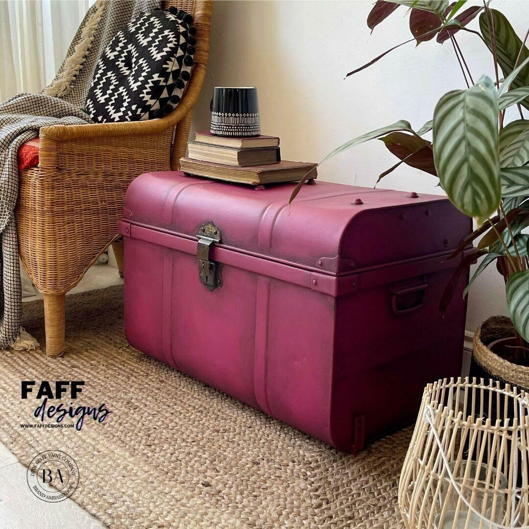 A magenta-pink vintage-style leather trunk chest with metal hardware, styled in a cozy room corner next to a wicker armchair, a potted plant, and a woven rug. The brand logo 'FAFF designs' is visible in the bottom left corner.