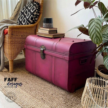 A magenta-pink vintage-style leather trunk chest with metal hardware, styled in a cozy room corner next to a wicker armchair, a potted plant, and a woven rug. The brand logo 'FAFF designs' is visible in the bottom left corner.