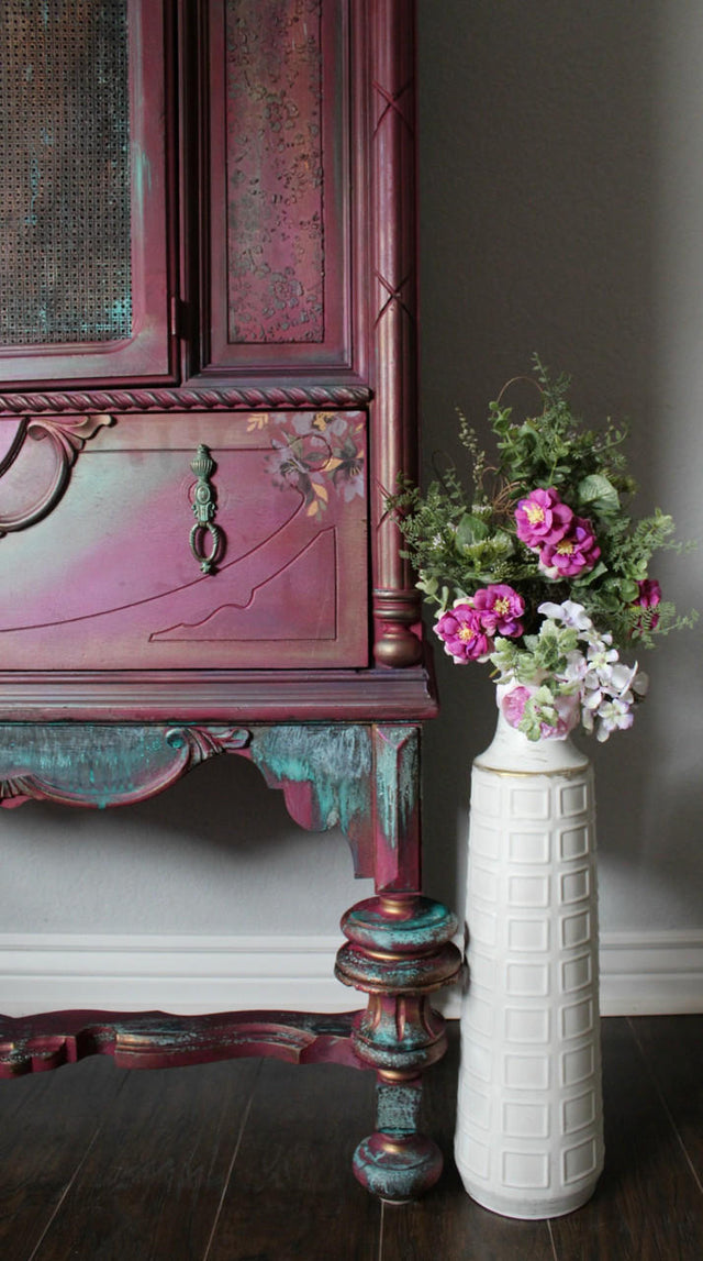 A close-up of an ornate vintage-style cabinet with distressed pink, purple, and teal paint, next to a tall white textured vase filled with pink and white flowers, set against a dark gray wall and dark wood floor.