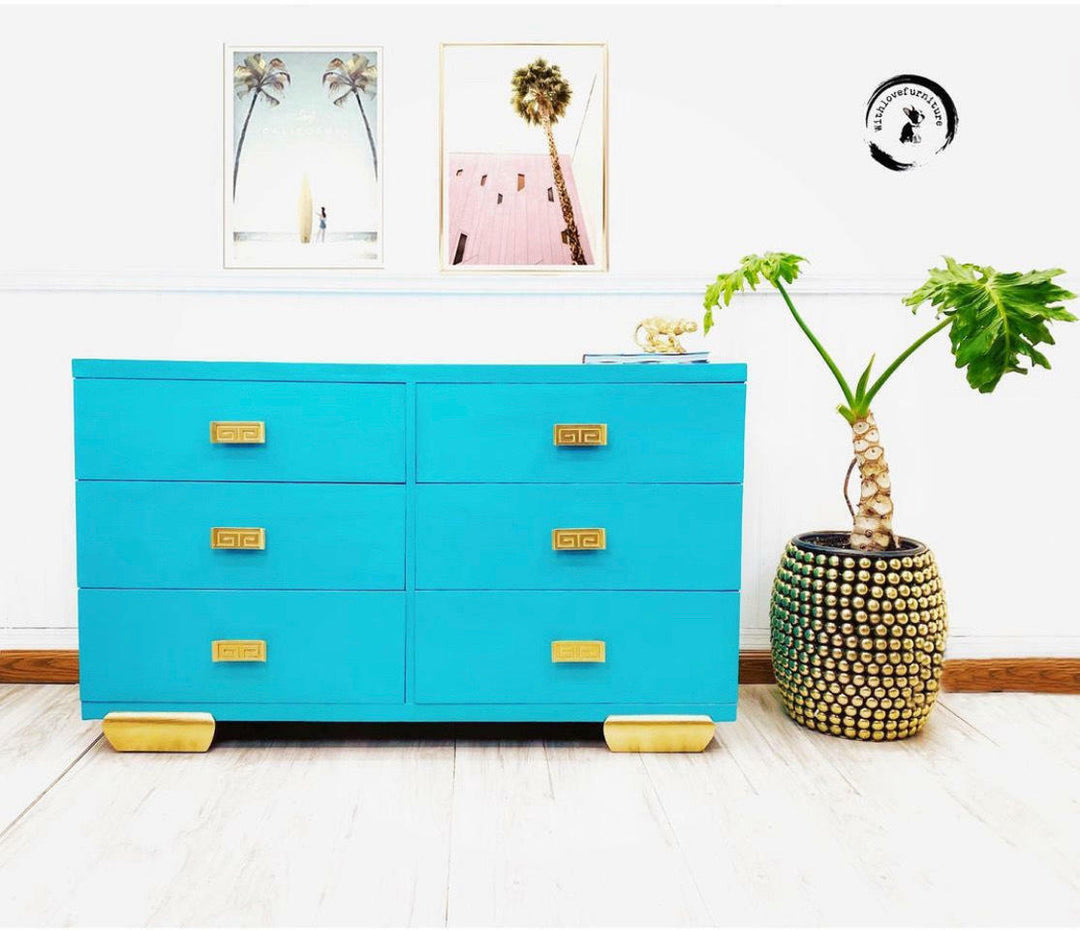 A bright turquoise six-drawer dresser with gold drawer pulls and gold feet, styled in a room next to a potted plant with a white wall and framed art in the background.
