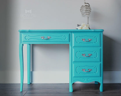 Turquoise desk with decorative handles against a gray wall