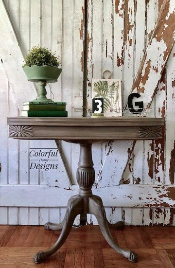 A distressed brown wooden console table with a turned pedestal leg and carved apron, styled with books, a green planter, and decorative letters against a rustic white wood paneled wall.