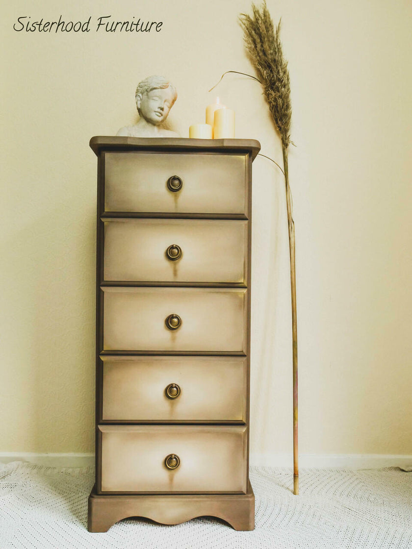 A vintage-style five-drawer chest of drawers with a distressed beige and brown finish, styled with a small bust, candles, and a dried pampas grass stem against a cream wall.