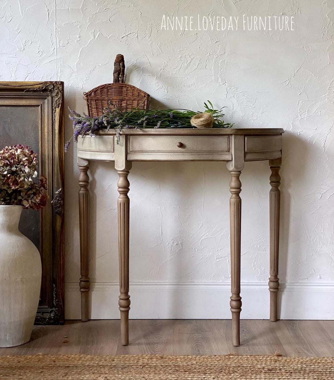 A distressed light beige console table with turned legs and a single drawer, styled with a wicker basket, dried flowers, and greenery against a textured white wall. A large framed picture and a cream vase with dried flowers sit to the left, and the text 'ANNIE.