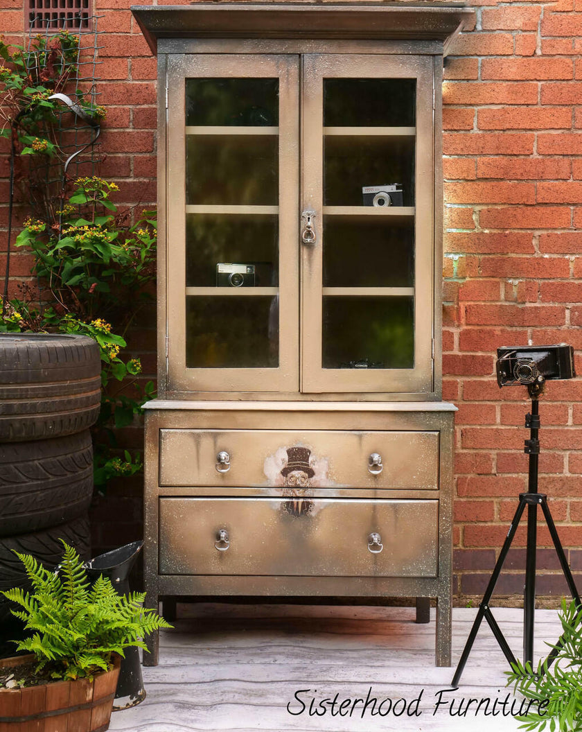 Vintage-style cabinet with glass doors and drawers against a brick wall, with 'Sisterhood Furniture' branding.