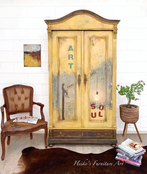 A vintage-style painted wooden cabinet stands against a white brick wall in a styled room. The cabinet has a distressed yellow and beige finish with painted text reading 'ART' and 'SOUL' and a dark silhouette figure.