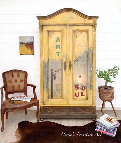 A vintage-style painted wooden cabinet stands against a white brick wall in a styled room. The cabinet has a distressed yellow and beige finish with painted text reading 'ART' and 'SOUL' and a dark silhouette figure.