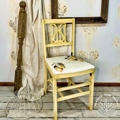 A light yellow wooden dining chair with a white upholstered seat featuring a gold pear design, set in a rustic room next to a wooden spindle and draped fabric.
