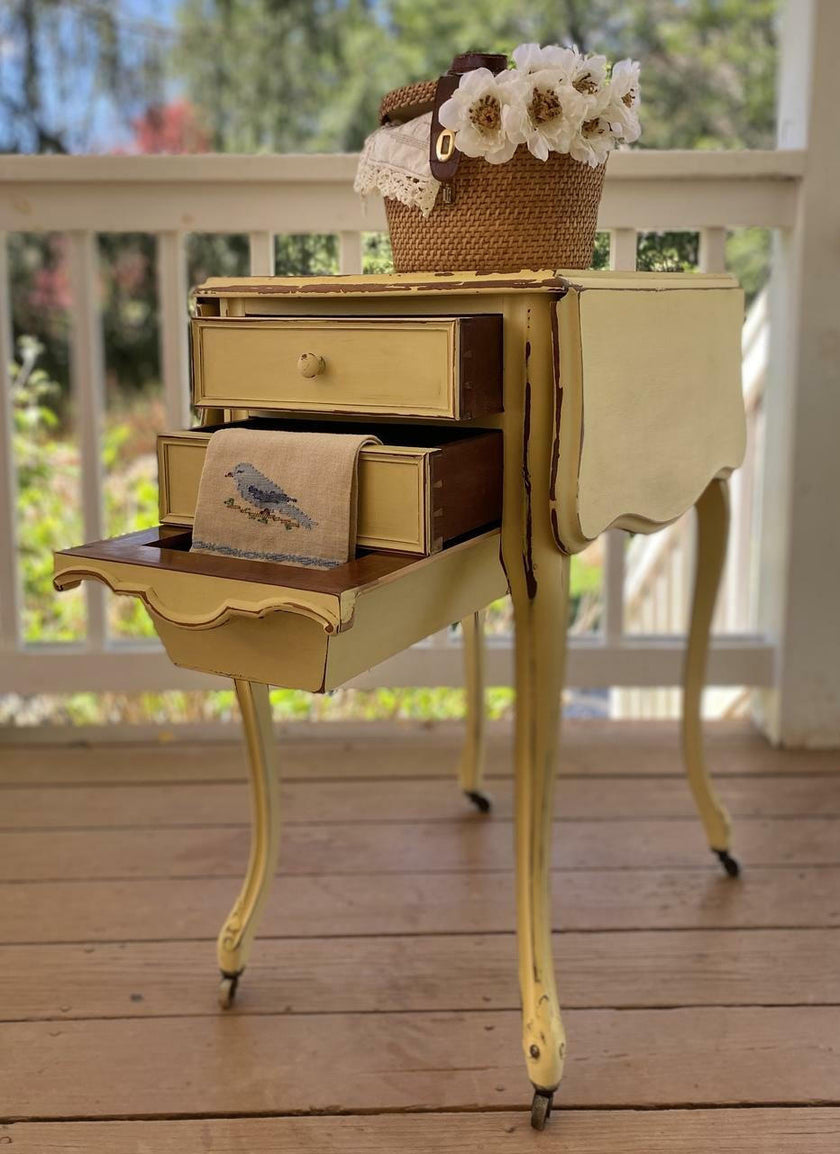 A vintage-style small table with a distressed yellow and brown painted finish, featuring a top drawer, a pull-out shelf with a bird print cloth, and a basket of white flowers on top, set on a wooden porch floor.