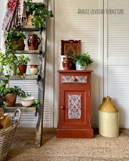 A rustic red wooden cabinet with decorative floral tile panels on the drawer and door, styled on a porch next to a ladder shelf with potted plants and a cream ceramic jug. The text 'ANNIE LOVEDAN FURNITURE' is visible in the top right.