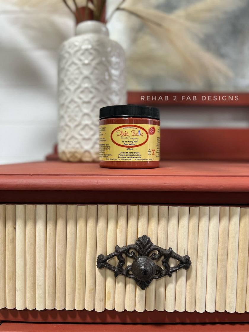 A jar of Dr. Sule's Professional Touch Enamel Paint in 'Rusty Nail' sits on top of a wooden chest of drawers with a ribbed beige front and a dark metal handle.