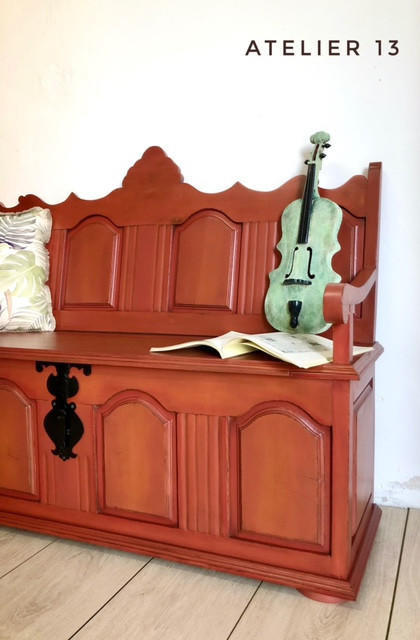 A reddish-brown wooden bench with decorative carved panels and a hinged front, styled in a room with a light-colored wall and wood floor. A green violin rests on the bench next to an open book, and a patterned pillow is visible on the left.