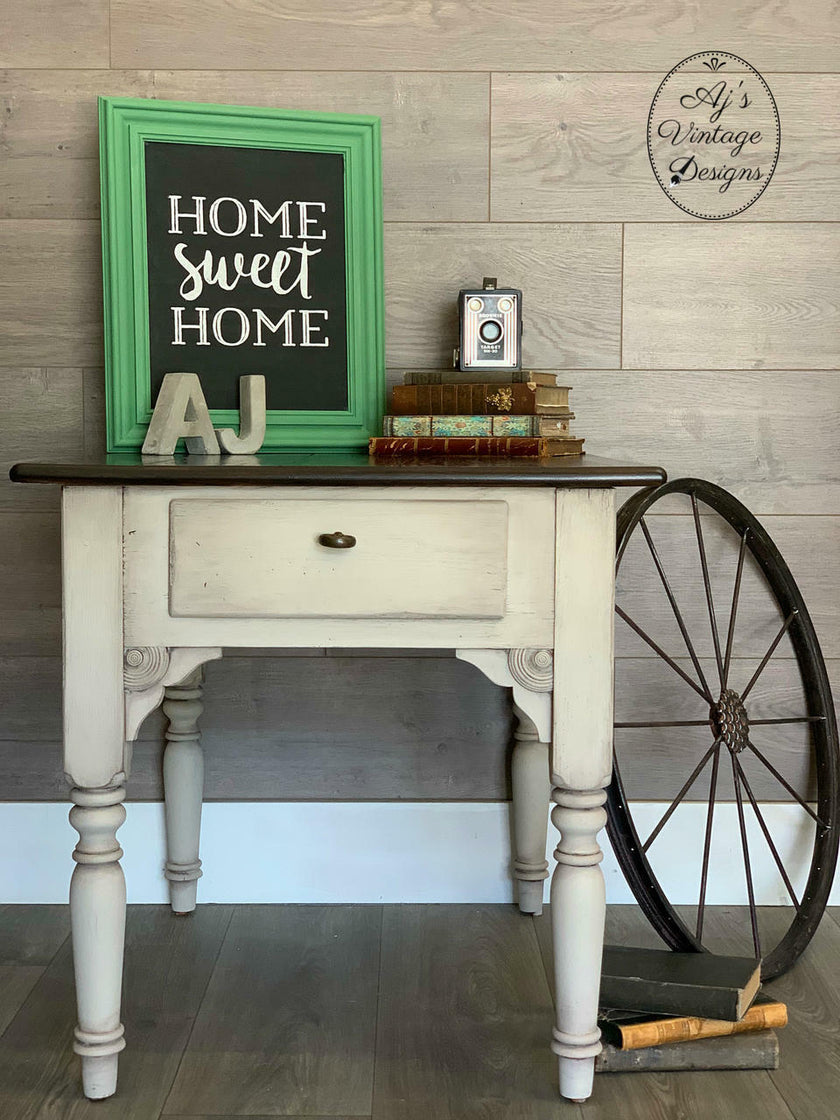 A distressed white wooden console table with a dark wood top, styled with a green framed chalkboard sign that reads 'HOME sweet HOME', vintage books, a small clock, and a large decorative wagon wheel leaning against a wood plank wall.