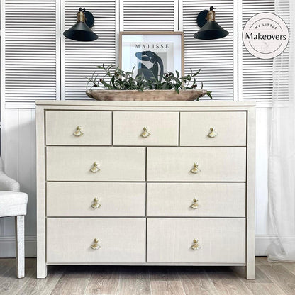 An off-white linen-textured 7-drawer dresser with gold-toned knobs, styled in a room setting with a decorative tray of greenery on top, black wall sconces, and a white shuttered wall background.