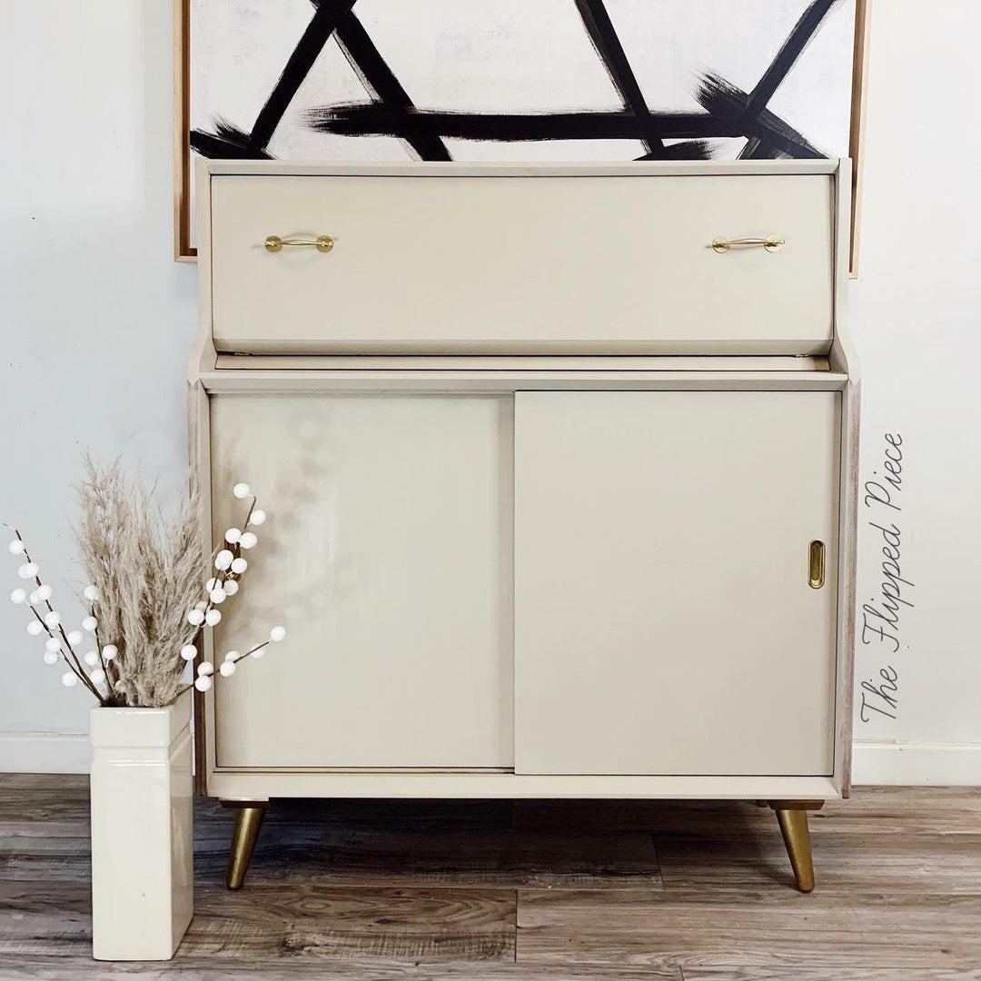 A mid-century modern cream-colored storage cabinet with a top drawer and two lower doors, accented with brass handles and legs. The cabinet is styled in a home setting on a wood floor next to a vase of dried grass, with a framed abstract print on the wall behind it.