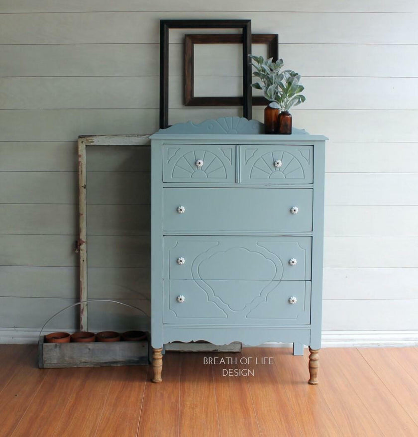 Blue dresser with decorative carvings on a wooden floor against a gray wall.