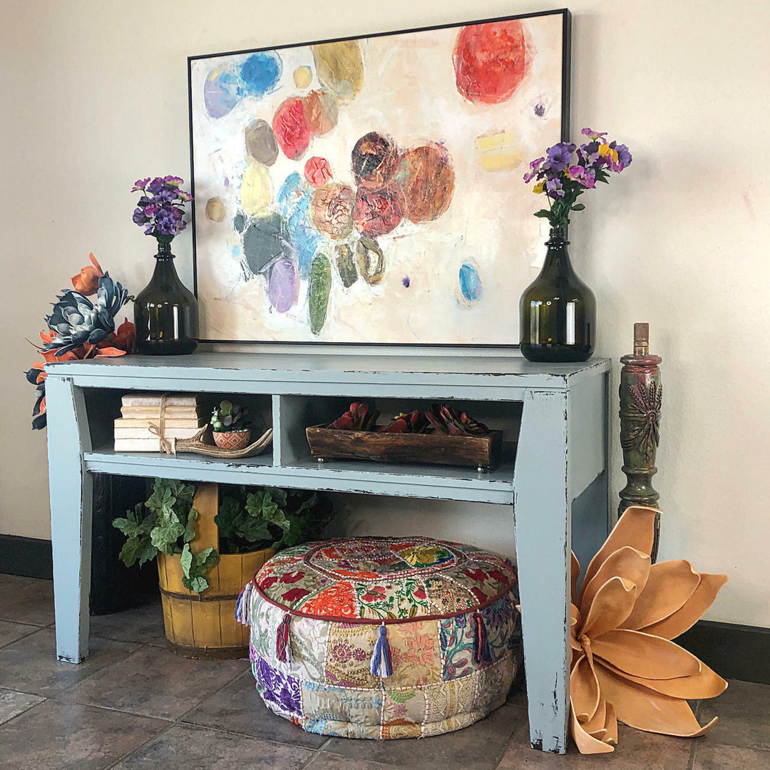 A styled interior scene showing a light blue console table with a large abstract painting above it, dark glass vases with purple flowers, books, baskets, potted plants, a patchwork ottoman, and decorative props against a plain wall.