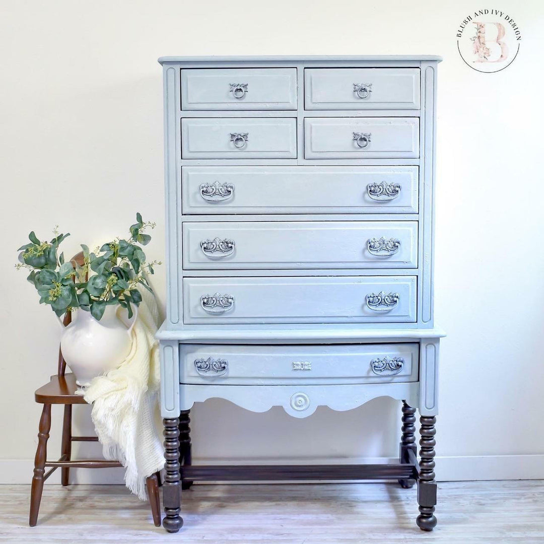 A tall, narrow vintage-style chest of drawers in a light blue-gray painted finish with ornate silver drawer pulls and dark turned legs, shown against a plain light wall with a small wooden stool and vase of greenery beside it.