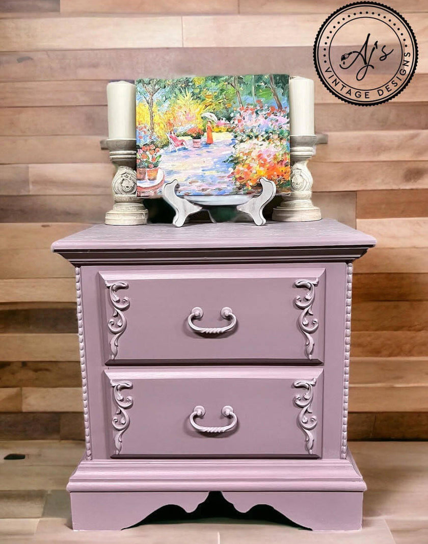 A lavender-painted two-drawer nightstand with ornate silver drawer pulls and decorative corner flourishes, styled with candlesticks and a colorful landscape painting against a wood plank wall.