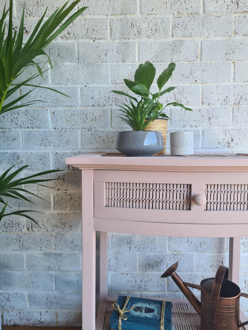 A light pink wooden console table with a woven rattan drawer front and single round knob, styled with a potted green plant, a small white cup, a blue book, and a metal watering can against a white brick wall.